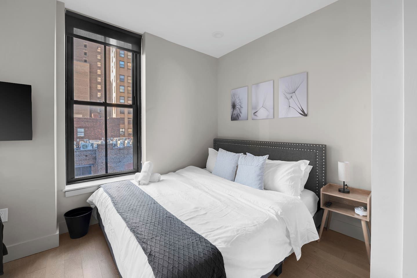 A bedroom with a large bed, a nightstand, a lamp, a window with a city view, and artwork on the wall.