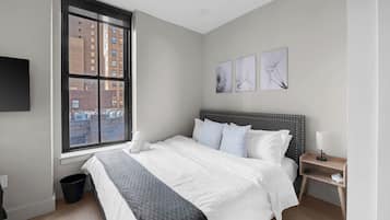 A bedroom with a large bed, a nightstand, a lamp, a window with a city view, and artwork on the wall.