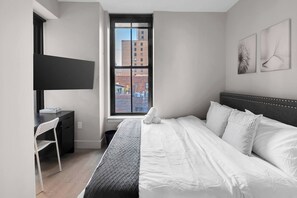 A modern bedroom with a bed, a desk, and a window with a city view. - Cozy Luxe 1BR –20 Min to NYC Rooftop & Gym Access (Newark)