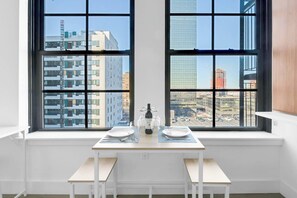 A dining area with a table set for two, a window with a city view, and a white interior. - Lux King 1BR 20 Min NYC 4 MIN to Prudential/Penn (Newark)