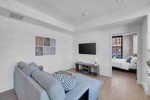 A modern living room with a grey sofa, a flat-screen TV mounted on the wall, and a bed with a window view. - Lux King 1BR 20 Min NYC 4 MIN to Prudential/Penn (Newark)