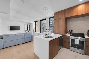 A modern kitchen with a built-in island, a dining area with a grey sofa, and a large window providing a view of the city. - Lux King 1BR 20 Min NYC 4 MIN to Prudential/Penn (Newark)