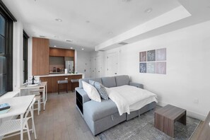 Eine moderne Wohnung mit einer Küchenzeile, einem grauen Sofa, einem kleinen Esstisch und einem weißen Bett.