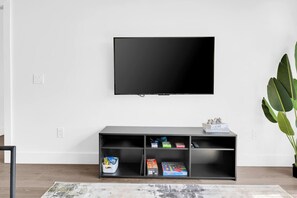 En moderne stue med et fladskærms-tv på væggen, et sort tv-møbel nedenunder og en potteplante til højre.