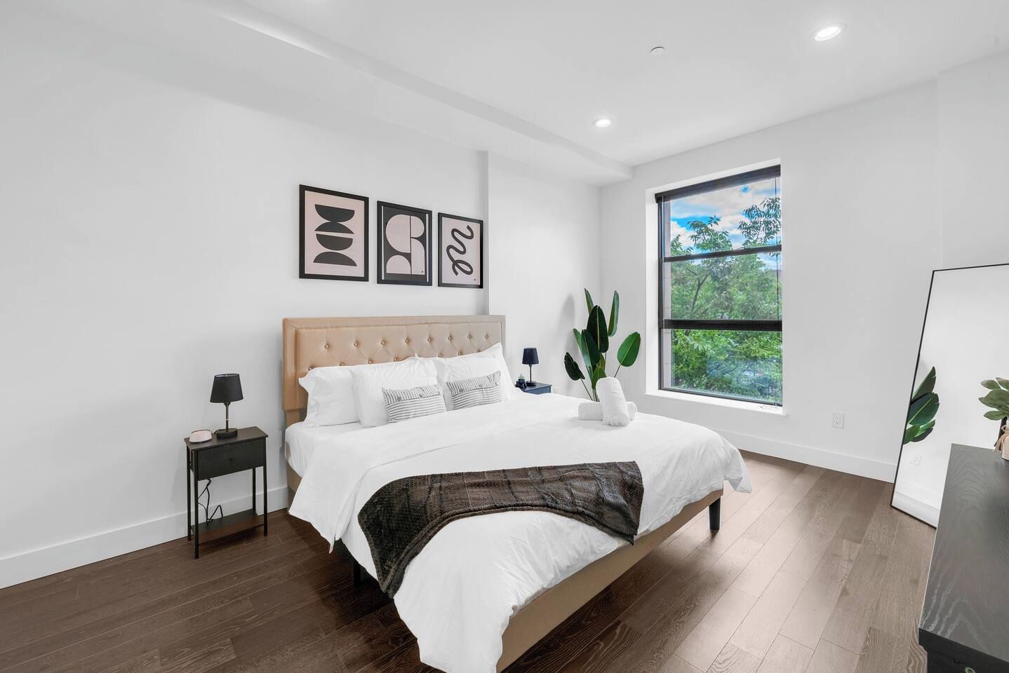 A modern bedroom with a large bed, a bedside table, a nightstand, and a window with a view of greenery.
