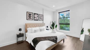 A modern bedroom with a large bed, a bedside table, a nightstand, and a window with a view of greenery.