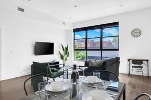 A modern living room with a dining table set for four, a sofa, a TV mounted on the wall, and a large window with a city view. - Central Stylish 1BR Free Parking Lux Fabulous King (Plainfield)
