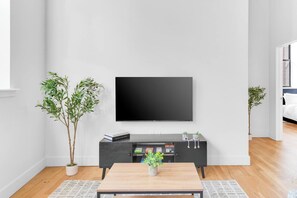 A modern living room with a flat-screen TV mounted on the wall, a dark wooden coffee table with a plant, and two potted plants on either side of the TV. - 2BR 20 min to NYC! Stylish Fun Cozy & Spotless (Newark)