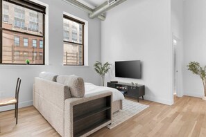 A modern bedroom with a sofa bed, a flat-screen TV, and a view of buildings through large windows. - 2BR 20 min to NYC! Stylish Fun Cozy & Spotless (Newark)