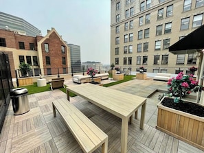 An outdoor patio with a wooden table and benches, surrounded by planters with flowers, and a trash bin. - 2BR 20 min to NYC! Stylish Fun Cozy & Spotless (Newark)
