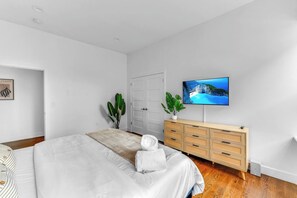 A modern bedroom with a bed, a TV, a dresser, and two potted plants. - 20 MIN to Manhattan! 2BR Luxurious Comfy & Spotless (Newark)