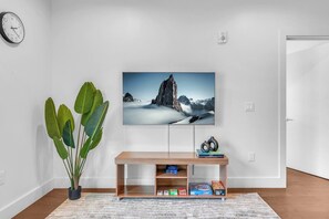 Ein modernes Wohnzimmer mit einem wandmontierten Flachbildfernseher, einem Holztischchen mit Büchern und Dekorationsobjekten, einer großen Topfpflanze und einer Wanduhr.