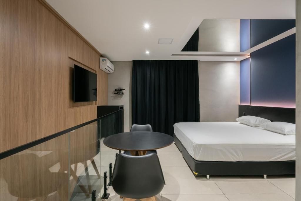 Habitación de hotel moderna con cama, una pequeña mesa con sillas, un televisor montado en la pared y una ventana con cortinas.