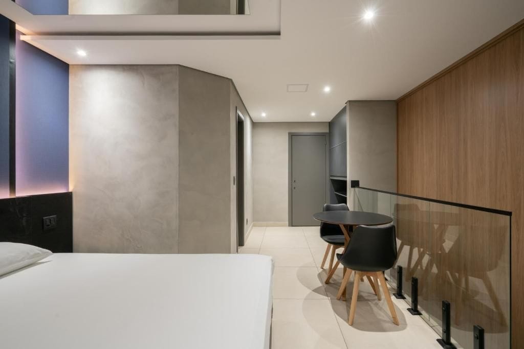 A modern hotel room with a bed, a small desk with chairs, and a glass partition.