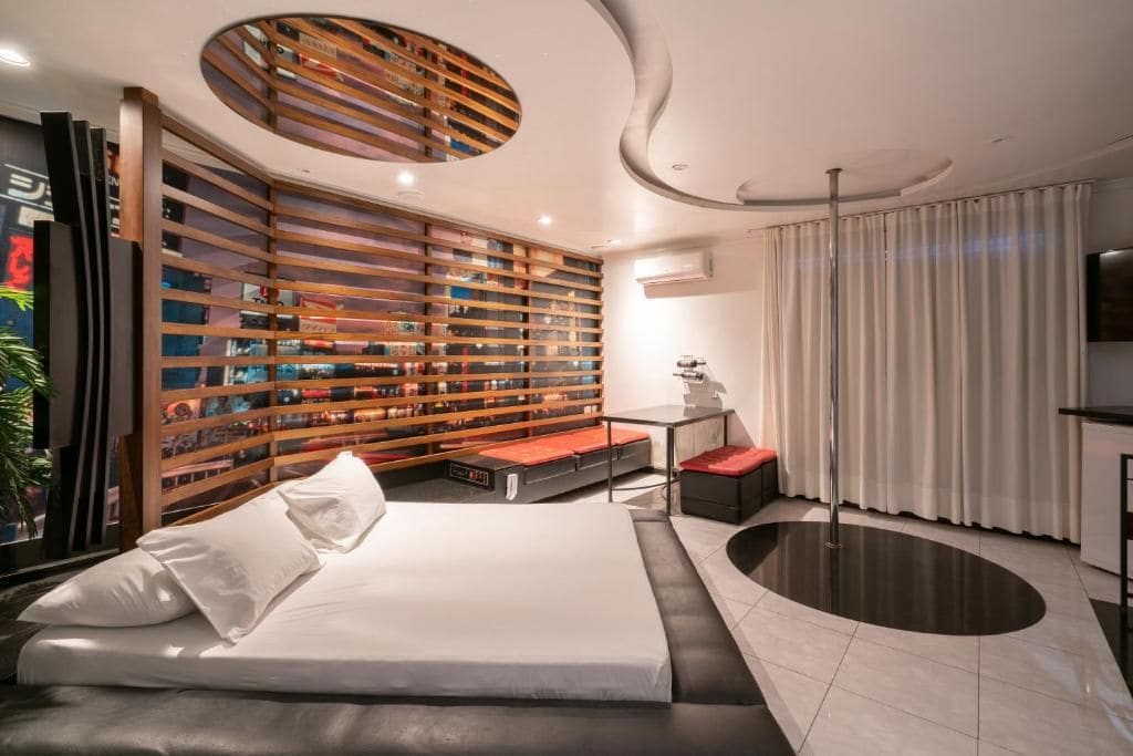Una habitación de hotel moderna con una cama grande, una estantería empotrada, una zona de estar circular y un ventanal con cortinas.
