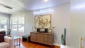 A living room with a herringbone-patterned wooden cabinet, a mounted artwork, and a neon sign reading 'Cowboy Cocktail Hour'. - Brazos Outpost ~ Ultimate Yard ~ 3BR/2BA (Bryan)