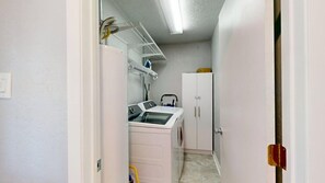 A laundry area with a washing machine, dryer, and shelves with cleaning supplies. - Brazos Outpost ~ Ultimate Yard ~ 3BR/2BA (Bryan)