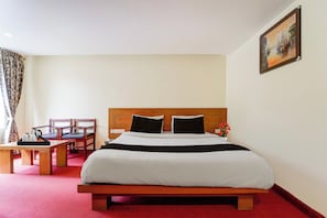 A hotel room with a bed, a wooden headboard, a painting on the wall, a desk with a chair, and a red carpet. - Townhouse Kodaikanal Hill Station (Kodaikanal)