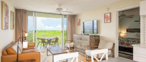 A room with a sofa, a table, a TV, a balcony with a view, and a bed with a floral bedspread.