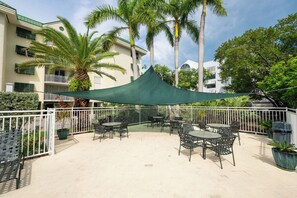 An outdoor patio area with tables and chairs, surrounded by a white fence, palm trees, and a building with balconies. - RUM CAY AT SUNRISE SUITES - 2 Bedroom / 2 Bathroom w/ Shared Pool, Spa, Parking (Key West)