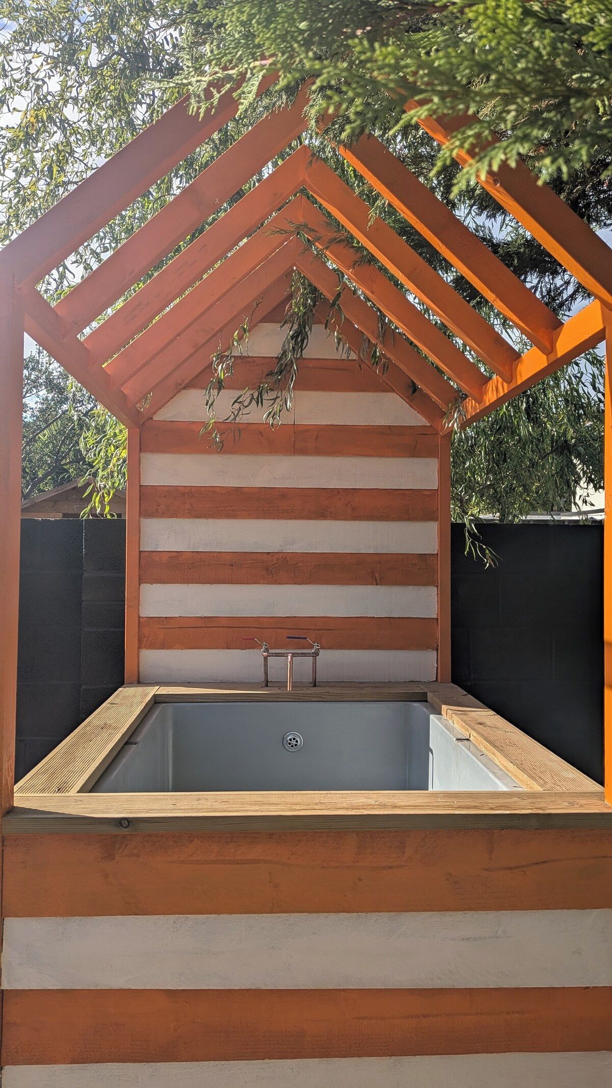 A hot tub with a wooden pergola overhead, surrounded by a black wall and trees.