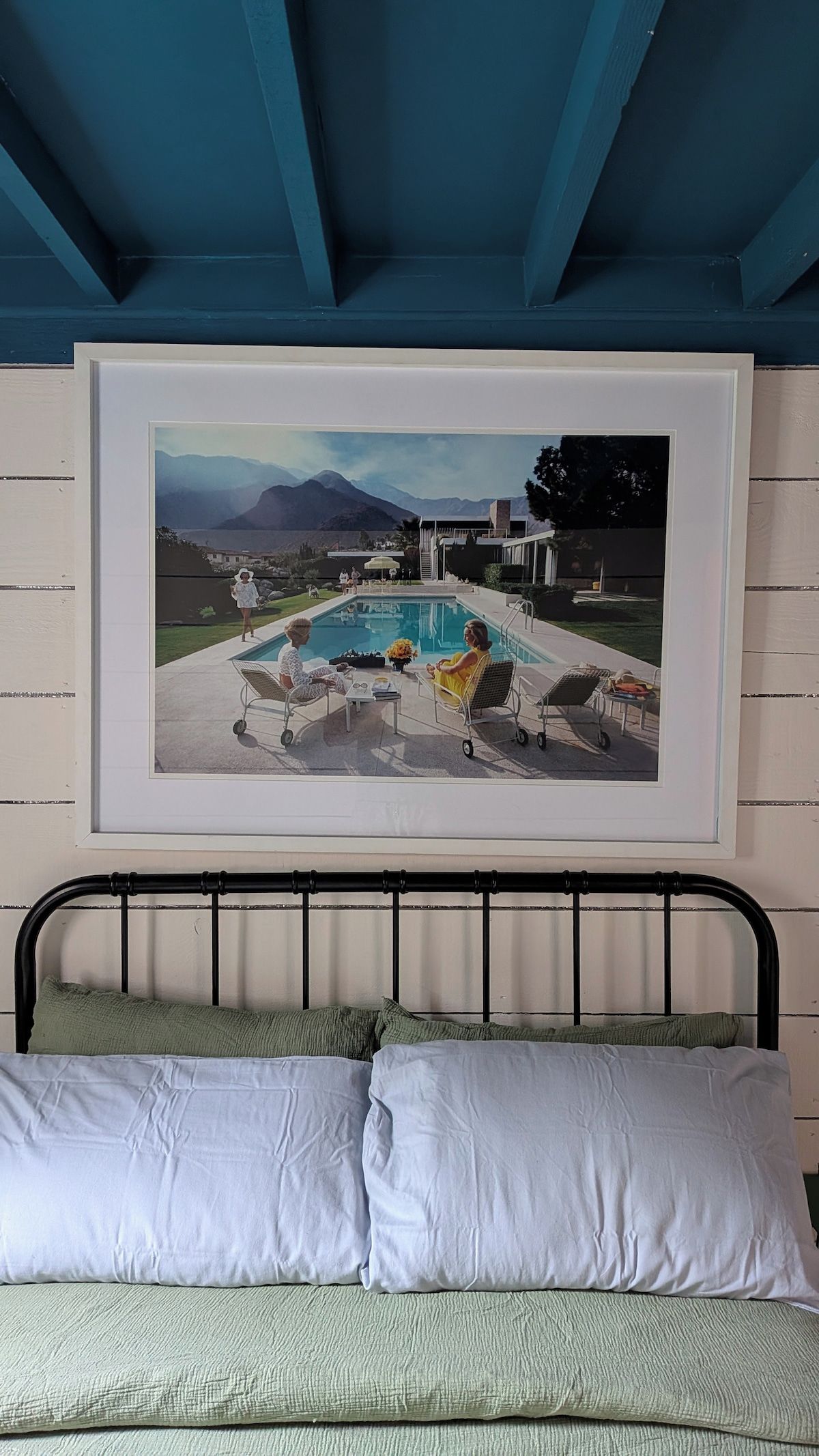 Fotografía enmarco de un área de piscina con montañas al fondo, una cama con ropa blanca y un techo azul.