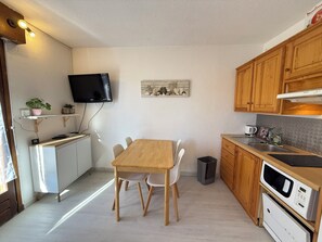A compact kitchen with a dining table, white cabinets, wooden cabinets, a microwave, and a television mounted on the wall. - Studio 4pers - terrasse - 300m des pistes (Guillaumes)