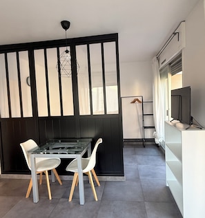 Una cocina moderna con una mesa de comedor de vidrio, dos sillas blancas, un panel negro y un televisor sobre un mueble blanco.