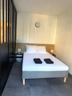 Habitación de hotel moderna con una cama grande, dos mesitas de noche, un reloj de pared redondo y vistas al exterior a través de una ventana con persianas verticales.