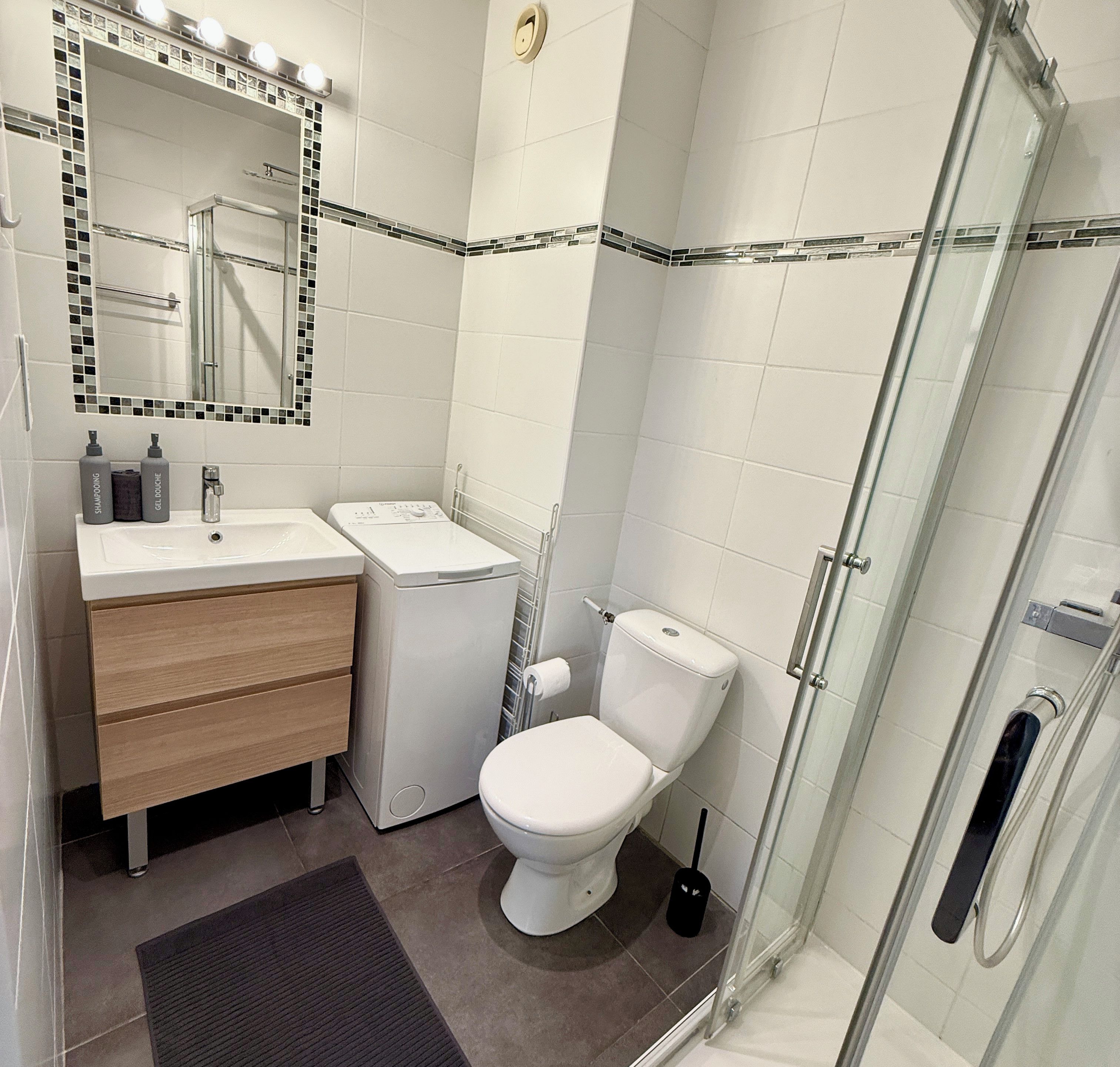 Une salle de bain moderne avec une douche à l’italienne en verre, des toilettes blanches, un meuble-lavabo avec un miroir et un petit tapis sur le sol.
