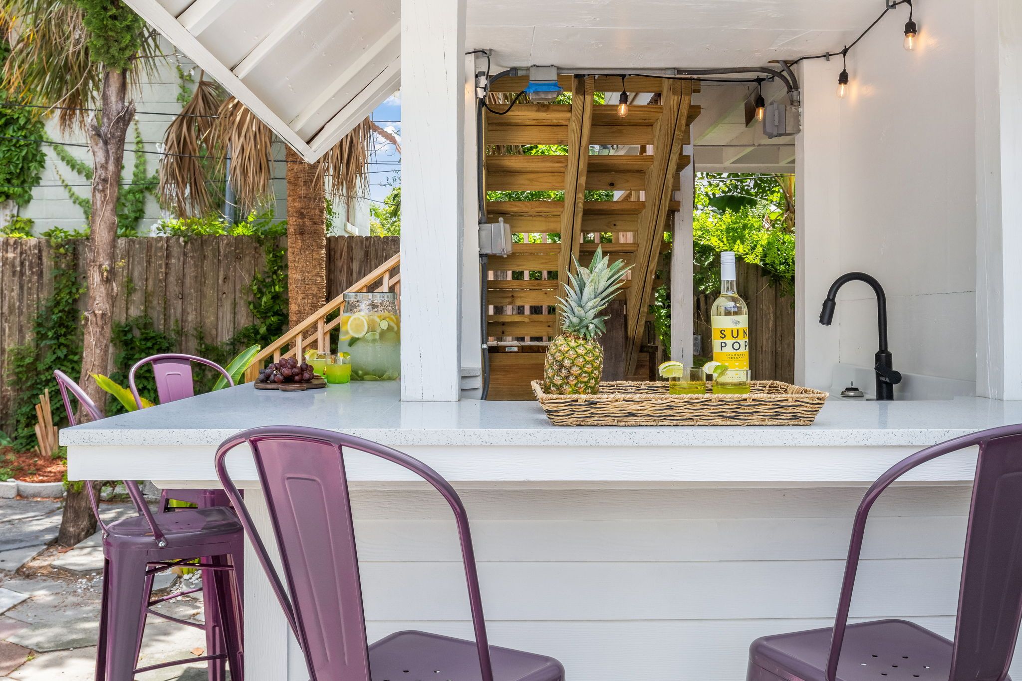 A bar area with a white countertop, purple chairs, a bottle of Sun Pop Lemonade, a pineapple, and a large glass pitcher.