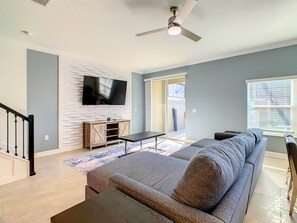 A living room with a grey sofa, a flat-screen TV mounted on a textured wall, a wooden entertainment unit, and a ceiling fan. - 5BRStorey Lake Resort | Private Pool | Near Disney (Kissimmee)