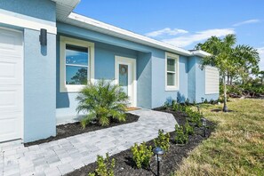 A residential building with a light blue exterior, a front door, two windows with shutters, a small garden with plants and a paved walkway. - Sunshine Retreat | NEW 4/2, King Bed, Near Beaches (Port Charlotte)