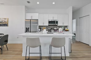 A modern kitchen with white cabinetry, stainless steel appliances, and a central island with bar stools. - Sunshine Retreat | NEW 4/2, King Bed, Near Beaches (Port Charlotte)