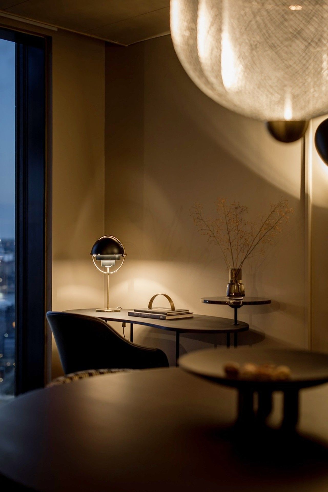 A modern interior with a dining area featuring a round table, a chair, a lamp, and a vase with branches.