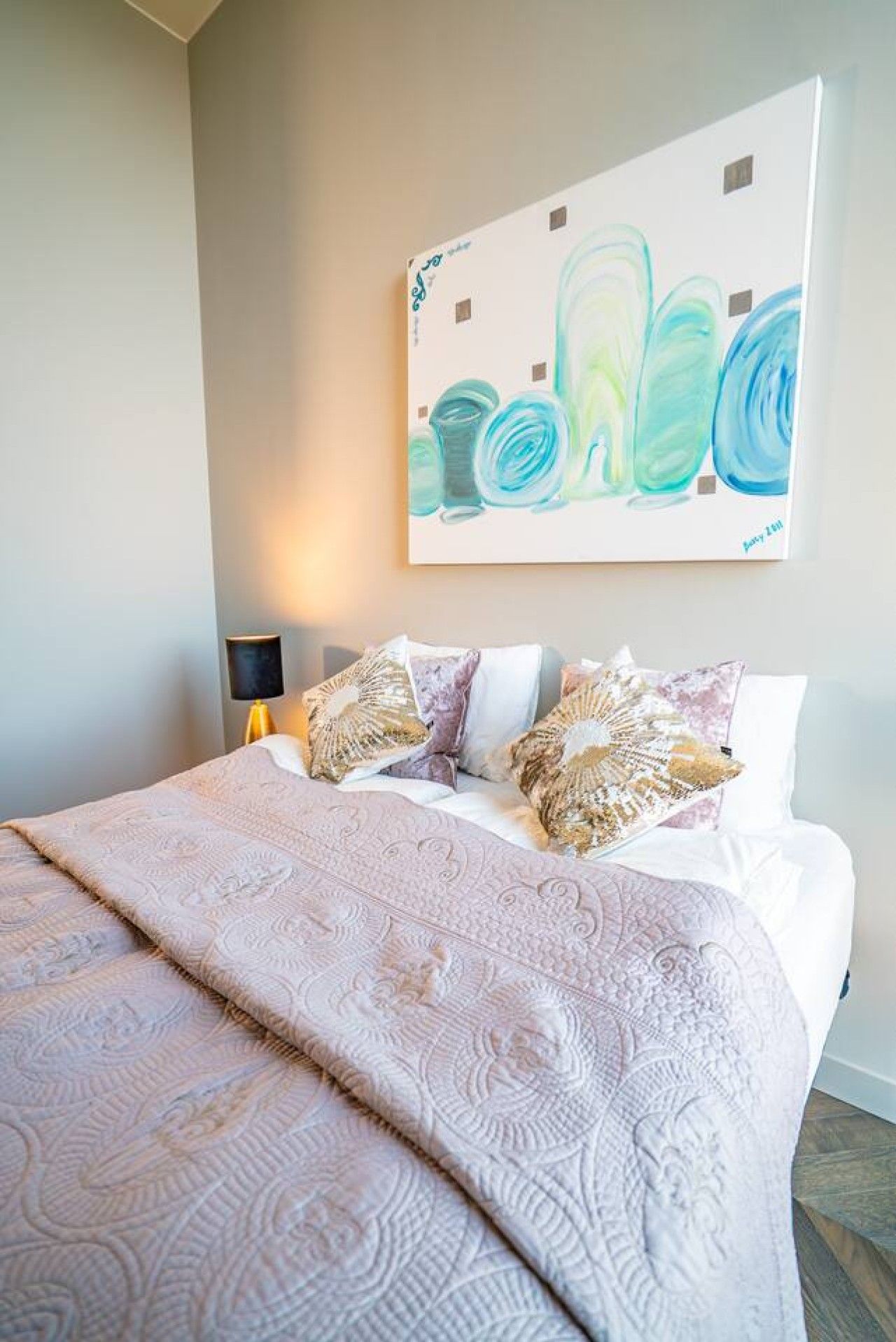 Una cama bien hecha con una colcha estampada y almohadas decorativas, un cuadro abstracto colgado en la pared y una lámpara de noche.