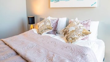 Una cama bien hecha con una colcha estampada y almohadas decorativas, un cuadro abstracto colgado en la pared y una lámpara de noche.