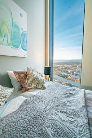 A bedroom with a large window, a bed with patterned bedding, and a city view.