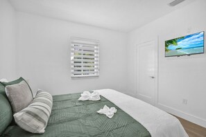 Ein Schlafzimmer mit Bett, Kissen, einem Fenster mit Jalousien, einer Tür und einem Fernseher an der Wand.