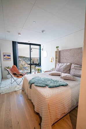 A modern bedroom with a large bed, a chair, a view through a window, and a balcony. - Karlatornet View Apartment (Gothenburg)