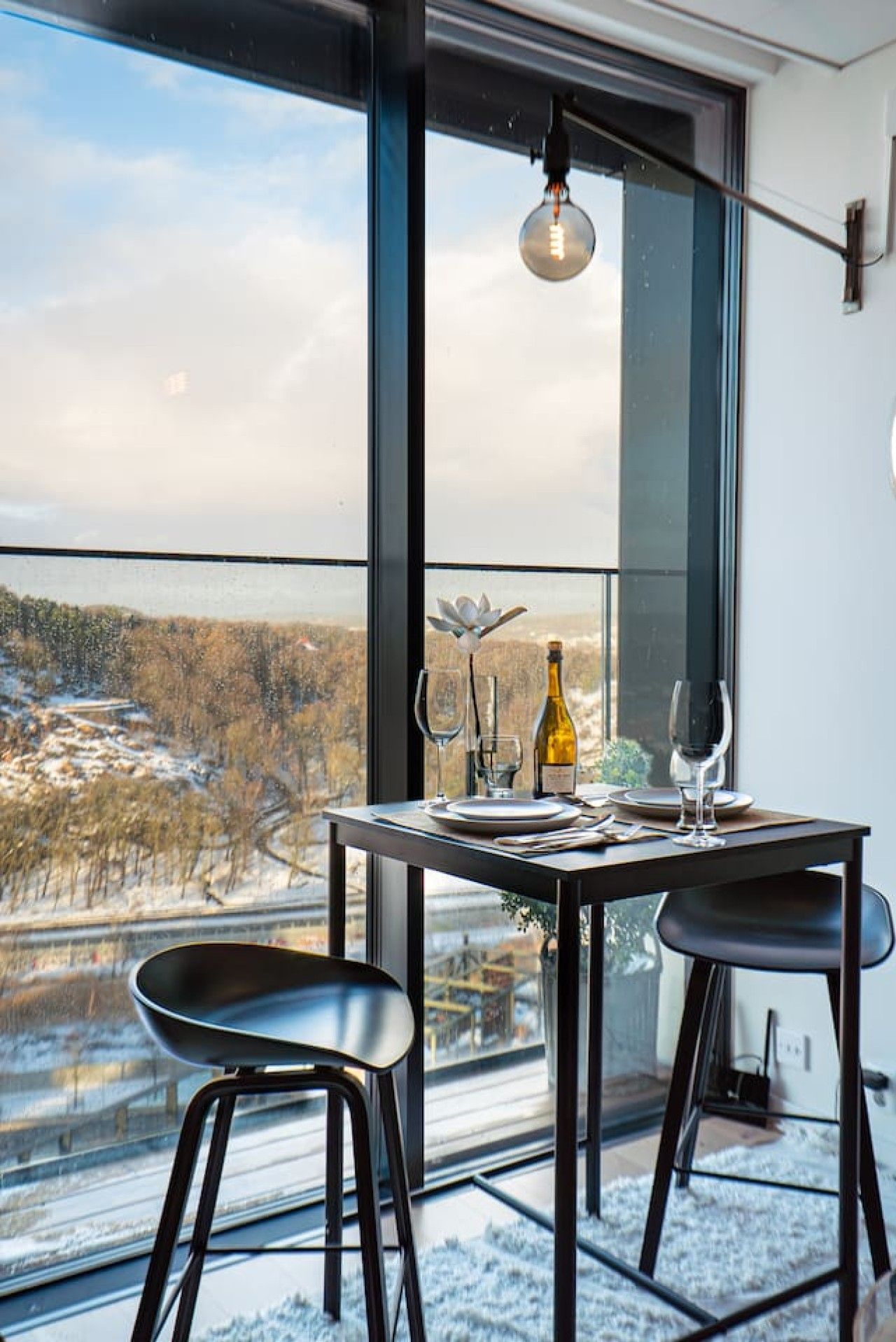 A dining area with a table set for two, a bottle of wine, and a view of a snowy landscape through large windows.