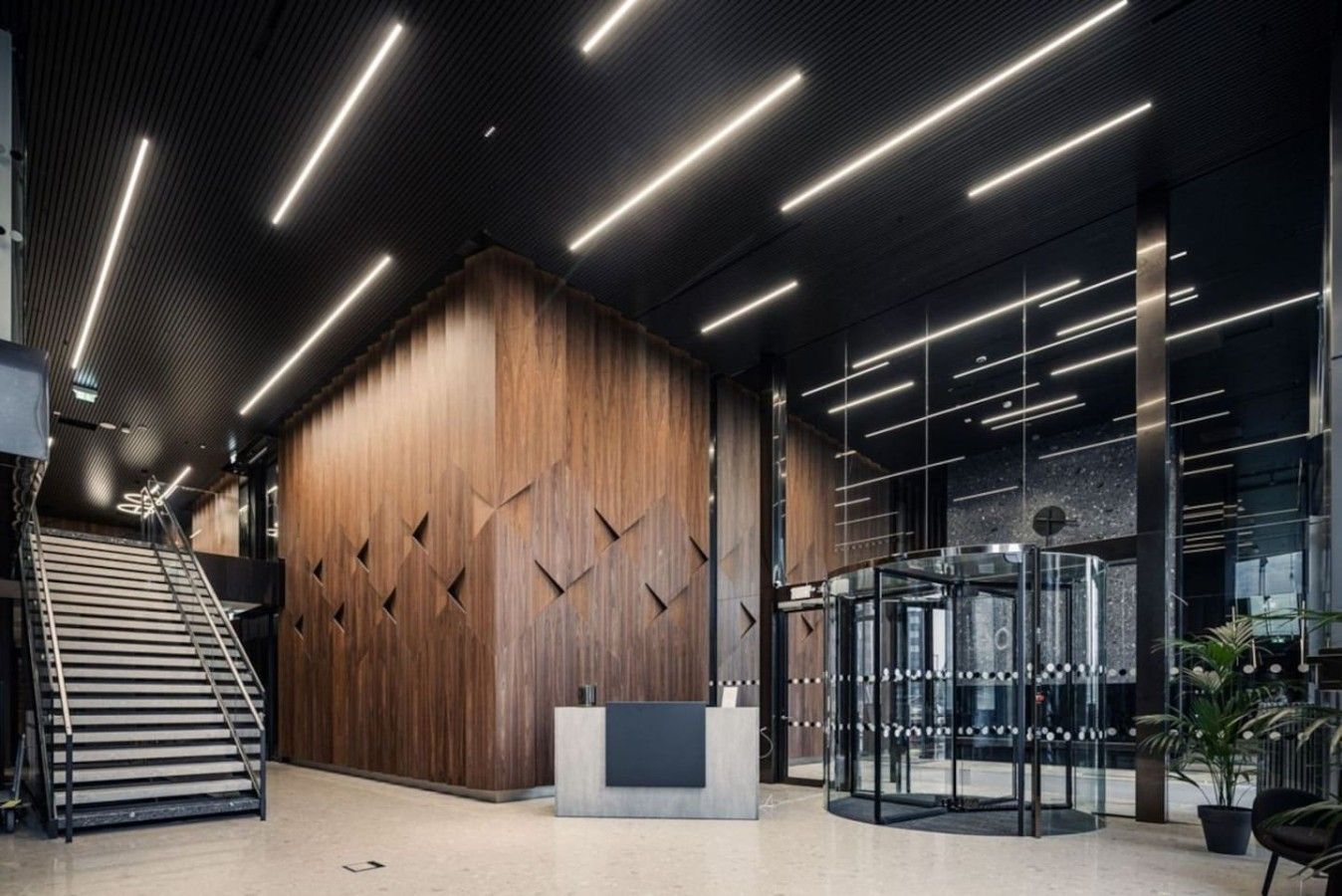 A modern lobby with a wooden reception desk, a glass elevator, and a staircase.