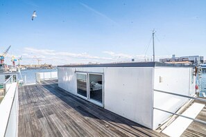 A deck with a small enclosed area, a view of the water, and a crane in the background.