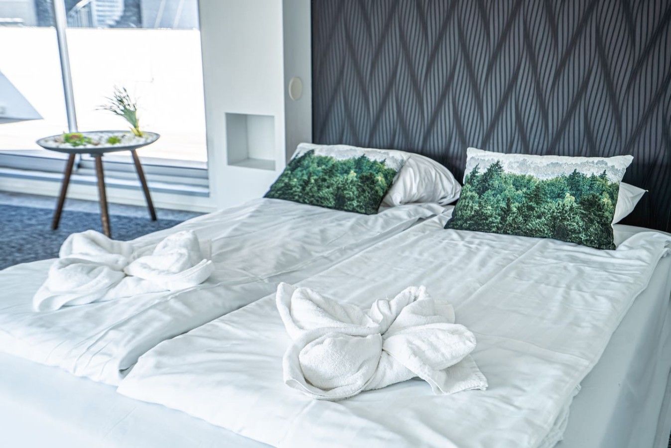 Hotellihuone, jossa on sänky, kaksi tyynyä ja pieni pöytä, jonka päällä on kasvi.