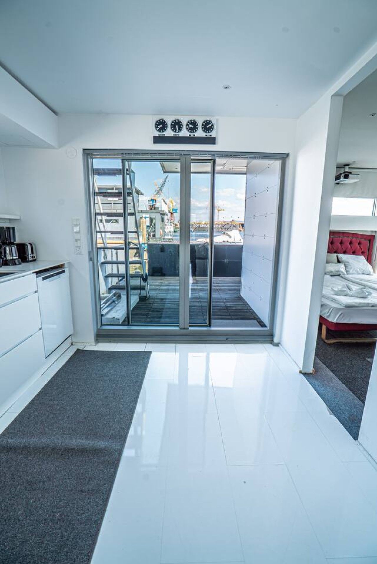 Un intérieur moderne avec une porte-fenêtre coulissante donnant sur un espace extérieur, une kitchenette équipée d’un lave-vaisselle et un lit avec une tête de lit rouge.