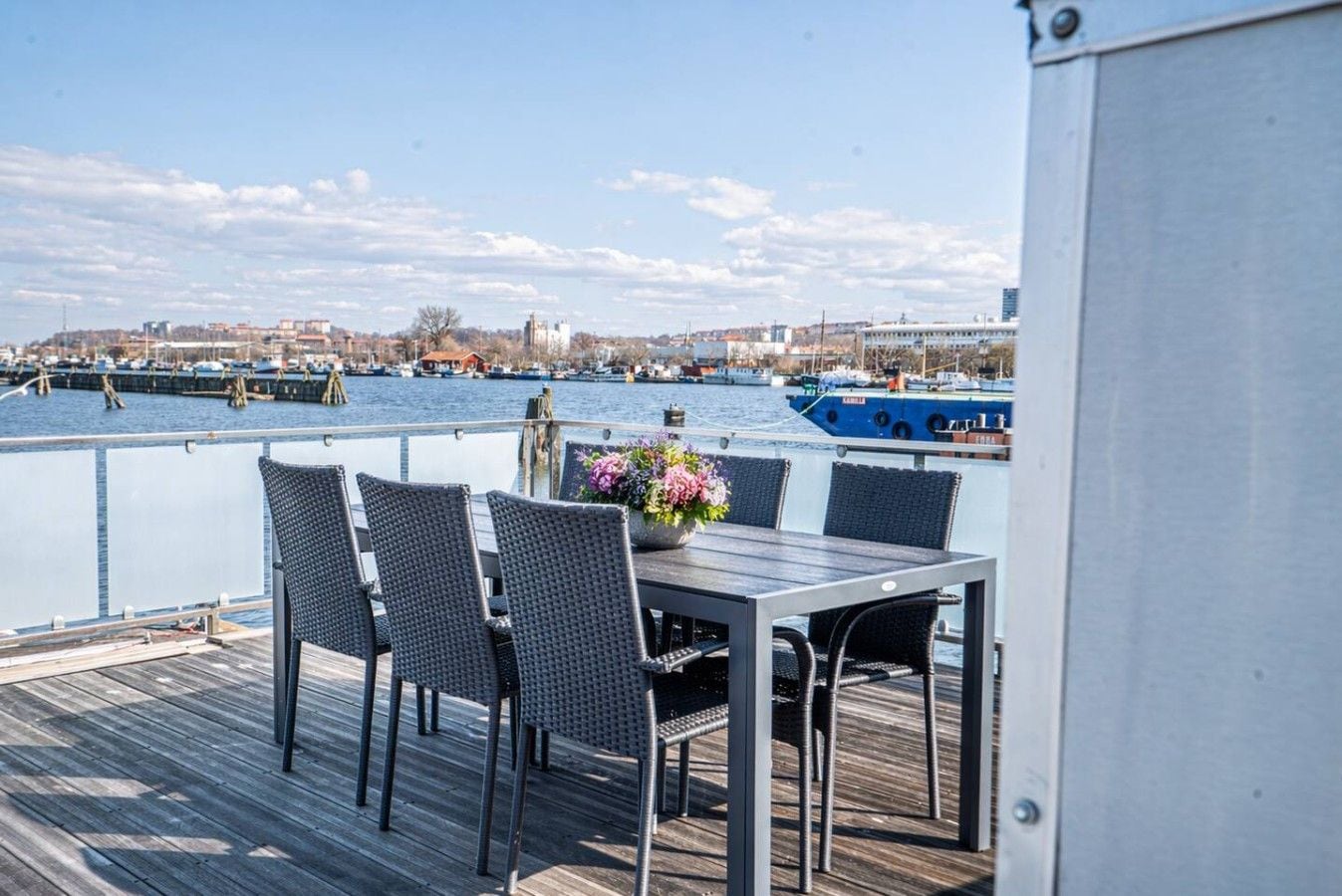 Une terrasse avec une table et des chaises, d’où l’on a vue sur une marina avec des bateaux et des bâtiments qui se dessinent à l’horizon.