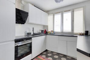 Une cuisine moderne avec des meubles blancs, un plan de travail noir, un four encastré et une fenêtre avec des stores.