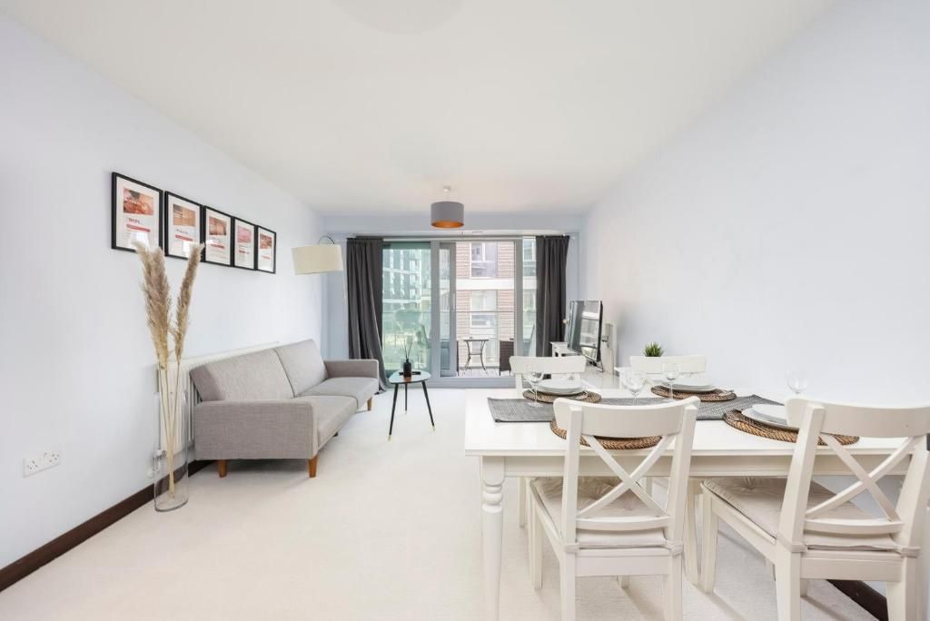 Een moderne woonkamer met een bank, eettafel en stoelen. Er is een groot raam met gordijnen, een lamp en ingelijste foto’s aan de muur.