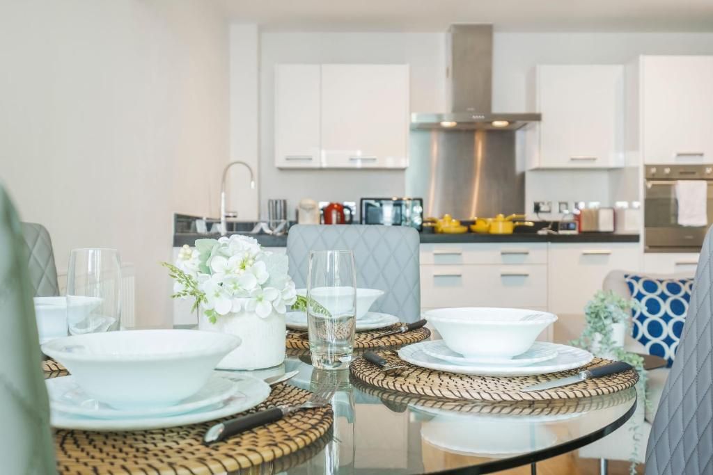 A modern kitchen with white cabinets, a stainless steel range hood, and a dining area with a glass table set with white plates and bowls.