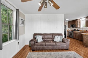 A living room with a brown leather sofa, a ceiling fan, and a view of the kitchen. - The Bayou Sanctuary | Peaceful Wooded Retreat (Ponchatoula)